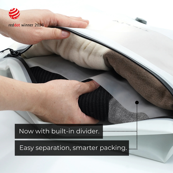 Hands lifting the grey divider inside a white Compak Medium to separate black knitwear from cream and brown clothing, captioned Now with built-in divider, easy separation, smarter packing