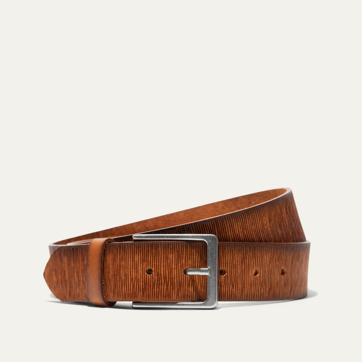 Will Leather Goods Wood Grain Embossed Belt coiled on a white background, tan leather with a fine wood-grain embossed texture and a brushed silver rectangular buckle.