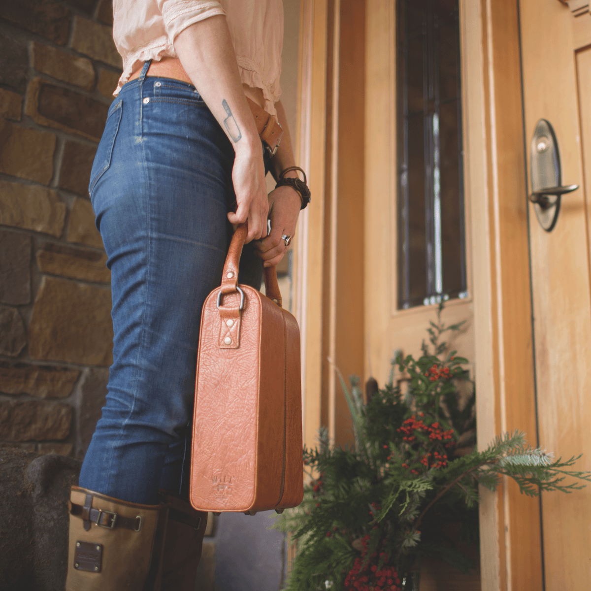 Lifestyle photo of a woman carrying the Will Leather Goods Hard Double Leather Wine Case in tan leather by the handle at a front doorway.