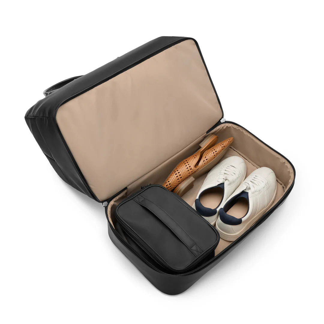 Open bottom shoe compartment of the Briggs & Riley Rhapsody Weekender Tote in black, holding a pair of white sneakers, a shoe horn, and a black zippered pouch