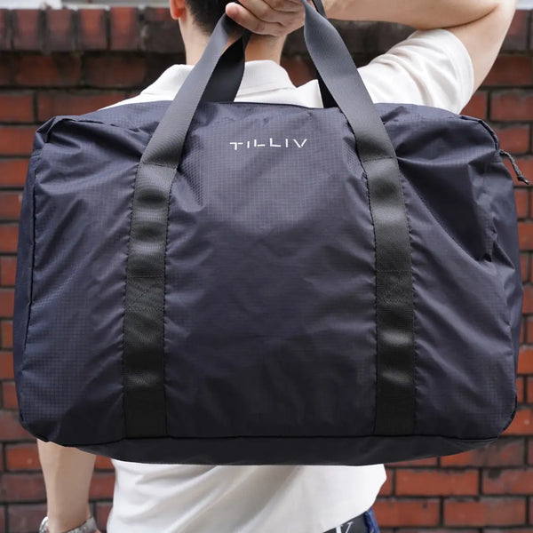 A person carrying a packed navy TILLIV Pakke duffel by its twin webbing handles against a brick wall