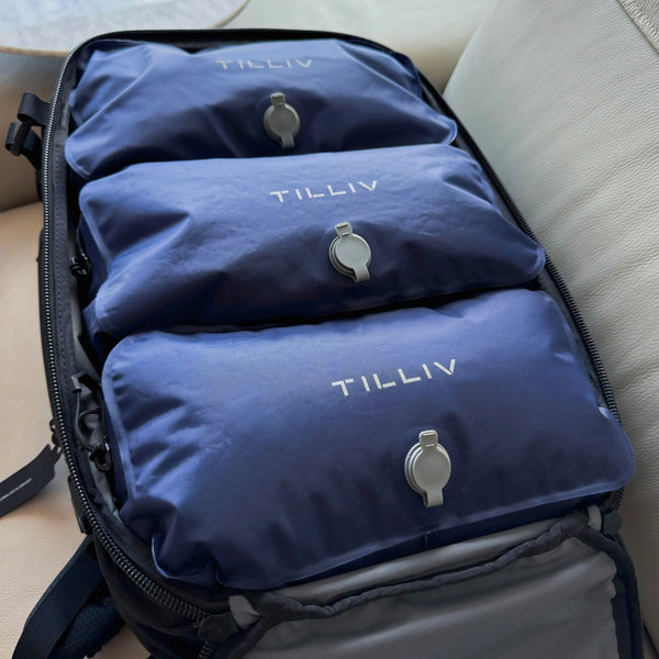 Three Midnight Blue TILLIV Stak packs stacked inside an open black travel backpack on a leather sofa