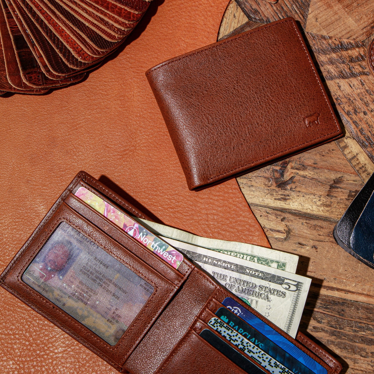 Flat lay lifestyle photo of the Will Leather Goods Classic Billfold Wallet in tan leather on a wood surface, filled with cash and cards, next to other leather wallets.