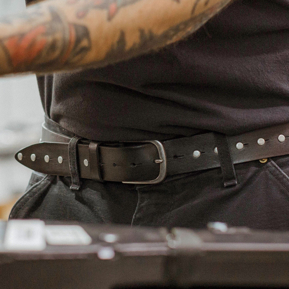 Lifestyle close-up of a tattooed man wearing the Will Leather Goods Single Studded Belt in black leather with a silver buckle, cinched over dark jeans.
