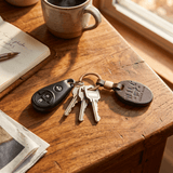 Dark brown leather oval key fob on a brass ring with a car key fob and three keys, resting on a wood table beside a coffee mug and notebook
