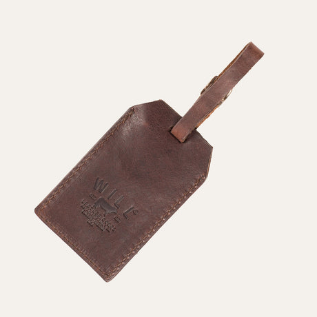 Dark brown leather luggage tag showing the embossed WILL Leather Goods logo side, with the buckled leather loop strap