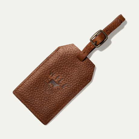 Cognac leather luggage tag shown at an angle, with the embossed WILL Leather Goods logo and a buckled leather loop strap