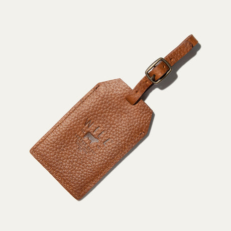 Tan leather luggage tag shown at an angle, with the embossed WILL Leather Goods logo and a buckled leather loop strap