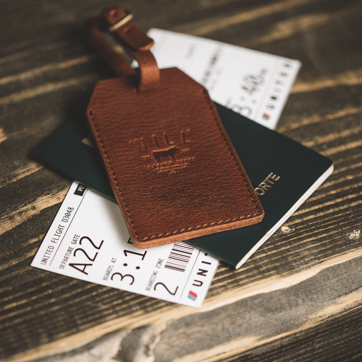 Lifestyle photo of a cognac leather luggage tag resting on a passport and boarding pass, with the strap buckled around luggage