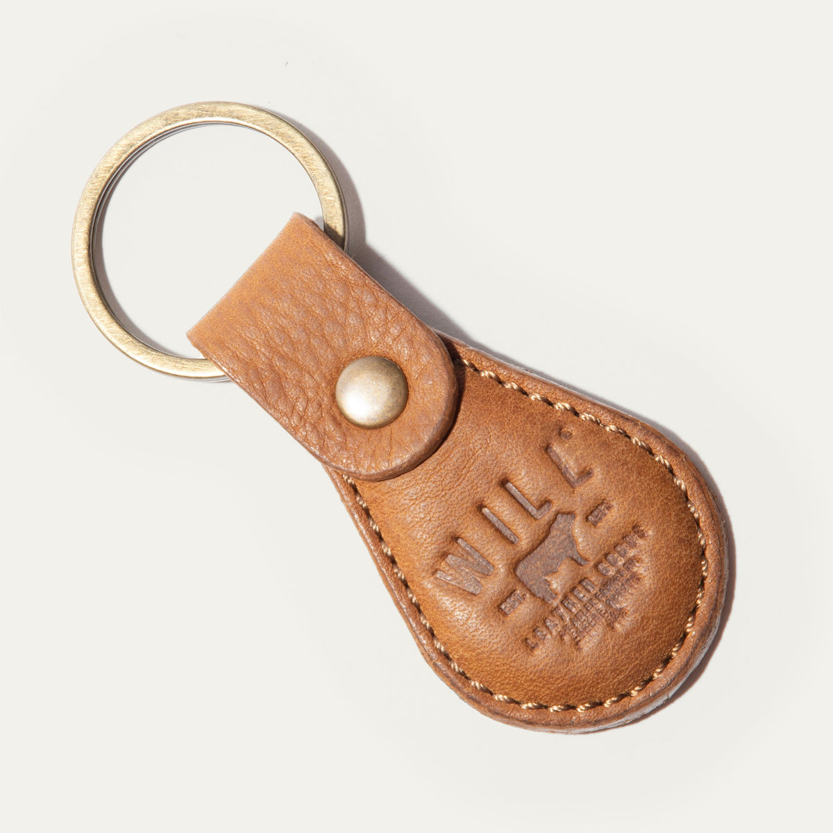 WILL Leather Goods logo keychain in cognac leather, teardrop-shaped fob with brass snap closure and keyring, embossed WILL Leather Goods medallion, on a white background.