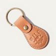 WILL Leather Goods logo keychain in cognac leather, angled close-up showing the brass snap closure and embossed WILL Leather Goods medallion, on a white background.