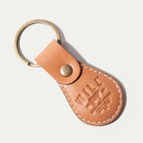 WILL Leather Goods logo keychain in cognac leather, angled close-up showing the brass snap closure and embossed WILL Leather Goods medallion, on a white background.