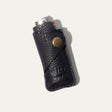 Black leather lighter cover with a BIC lighter tucked inside, angled view showing the brass snap closure and WILL Leather Goods embossed logo