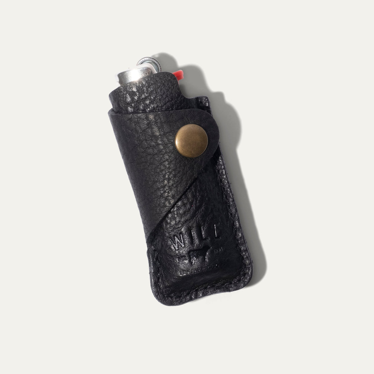 Black leather lighter cover with a BIC lighter tucked inside, angled view showing the brass snap closure and WILL Leather Goods embossed logo