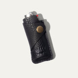 Black leather lighter cover with a BIC lighter tucked inside, angled view showing the brass snap closure and WILL Leather Goods embossed logo