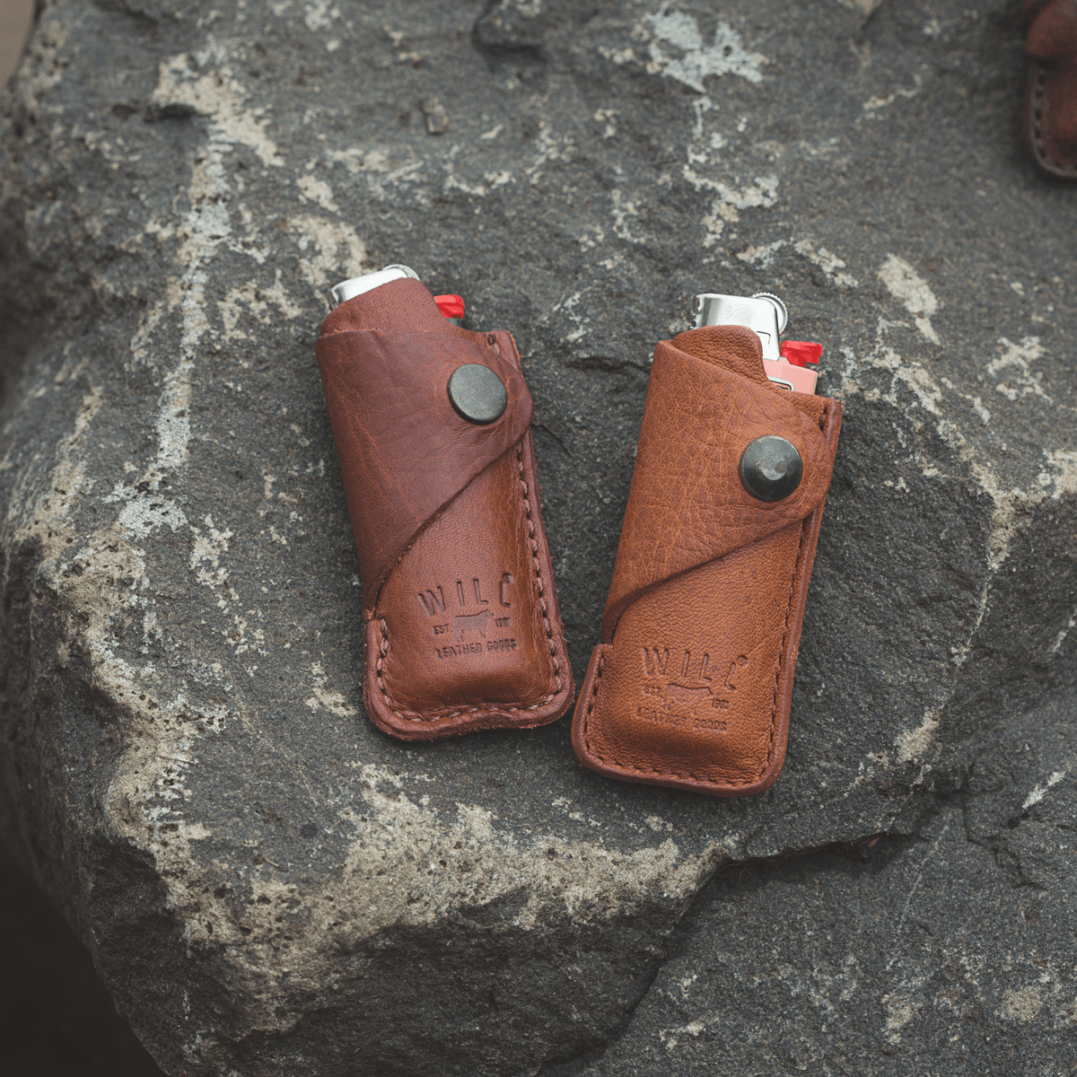 Two leather lighter covers, one oxblood and one tan, each holding a lighter, resting on a stone surface
