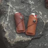 Two leather lighter covers, one oxblood and one tan, each holding a lighter, resting on a stone surface