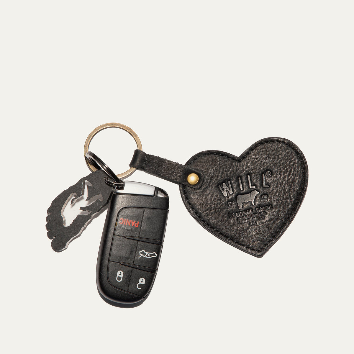Will Leather Goods Heart Keychain in black pebbled leather with embossed WILL logo, attached by a brass ring and snap loop to a car remote key and a small foot-shaped bottle opener, on a white background.