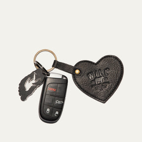Will Leather Goods Heart Keychain in black pebbled leather with embossed WILL logo, attached by a brass ring and snap loop to a car remote key and a small foot-shaped bottle opener, on a white background.