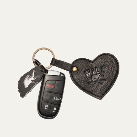 Will Leather Goods Heart Keychain in black pebbled leather with embossed WILL logo, attached by a brass ring and snap loop to a car remote key and a small foot-shaped bottle opener, on a white background.