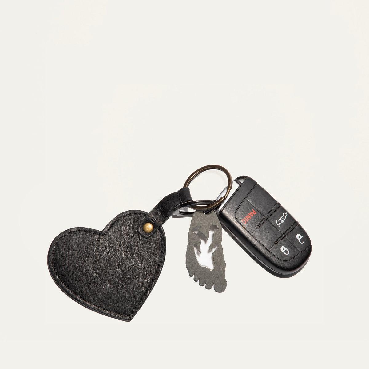 Overhead view of the black leather Heart Keychain with embossed WILL Leather Goods logo, clipped to a car key fob and foot-shaped bottle opener charm on a white background.