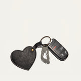 Overhead view of the black leather Heart Keychain with embossed WILL Leather Goods logo, clipped to a car key fob and foot-shaped bottle opener charm on a white background.