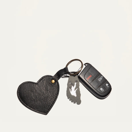 Overhead view of the black leather Heart Keychain with embossed WILL Leather Goods logo, clipped to a car key fob and foot-shaped bottle opener charm on a white background.