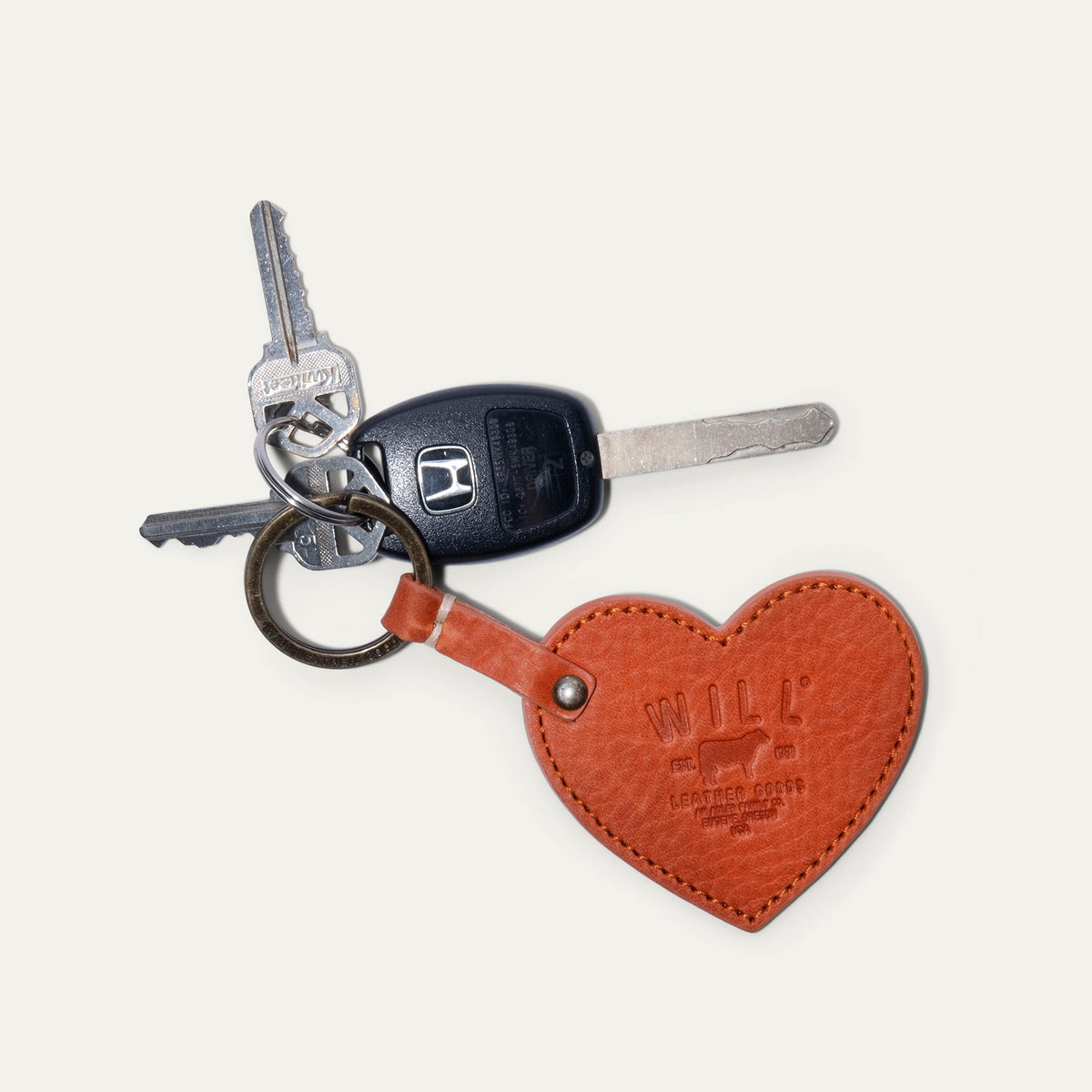Will Leather Goods Heart Keychain in cognac leather with embossed WILL logo, attached by a brass ring to a Honda car key and two household keys, on a white background.