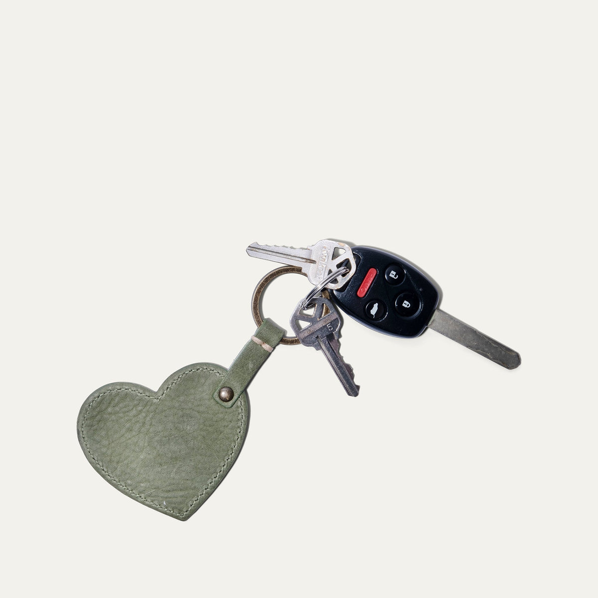 Overhead view of the sage green leather Heart Keychain with embossed WILL Leather Goods logo, attached to a Honda car key and two household keys on a white background.
