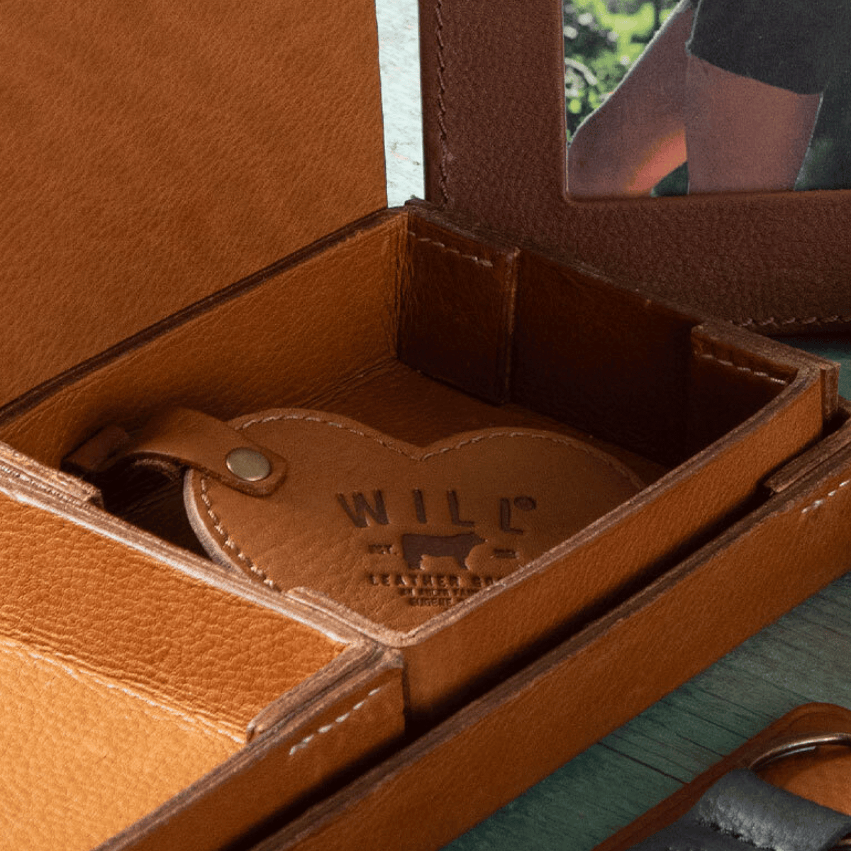 Close-up lifestyle photo of a tan leather Heart Keychain with embossed WILL Leather Goods logo tucked inside an open tan leather valet tray, with a lifestyle photo of a person outdoors visible in the background.