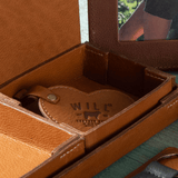 Close-up lifestyle photo of a tan leather Heart Keychain with embossed WILL Leather Goods logo tucked inside an open tan leather valet tray, with a lifestyle photo of a person outdoors visible in the background.