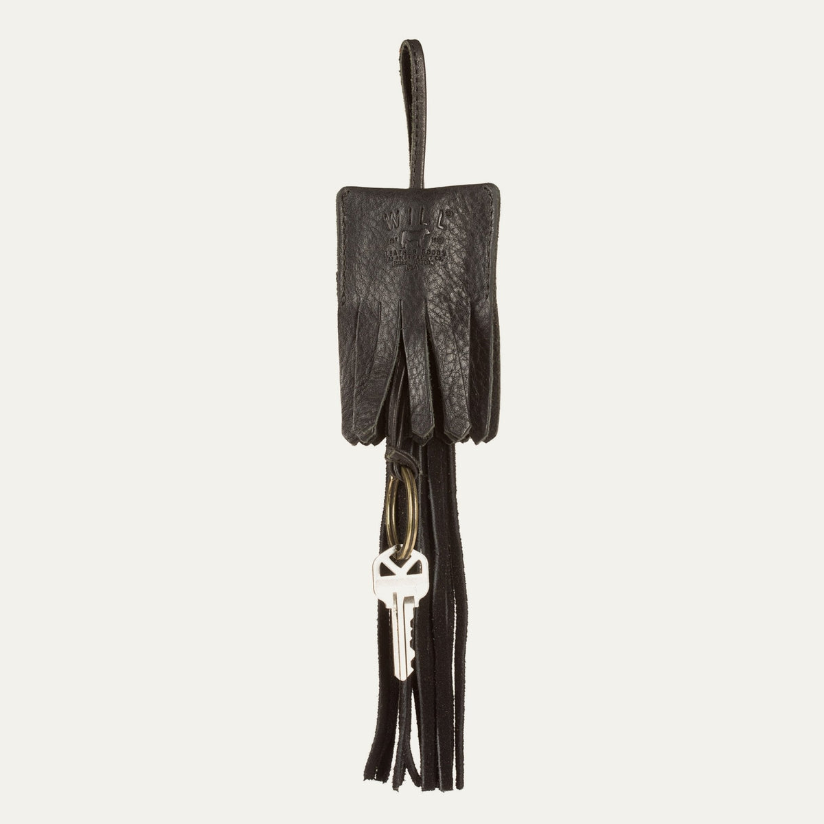 Close-up lifestyle photo of a black leather WILL Leather Goods tassel key fob with a single house key attached to its brass ring, against a white background.