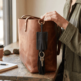 Lifestyle photo of a black leather WILL Leather Goods tassel key fob clipped to the handle of a cognac leather tote bag, with keys hanging from its brass ring.