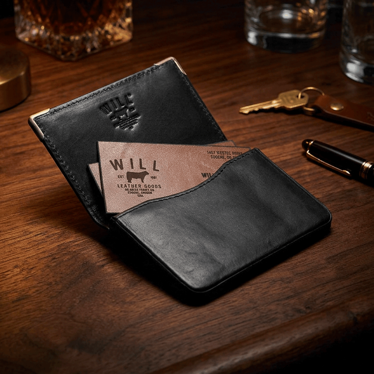 Lifestyle photo of the black card holder open on a wood desk with Will Leather Goods business cards, keys, and a pen