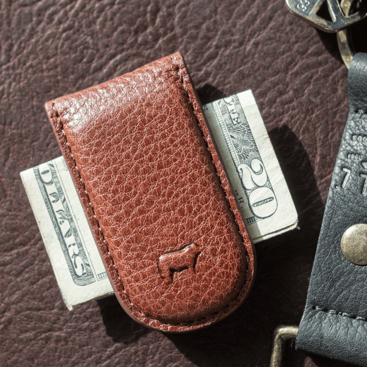 Close-up lifestyle photo of the tan leather Classic Money Clip holding folded bills, resting on a dark brown leather surface next to a keychain.