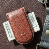 Close-up lifestyle photo of the tan leather Classic Money Clip holding folded bills, resting on a dark brown leather surface next to a keychain.