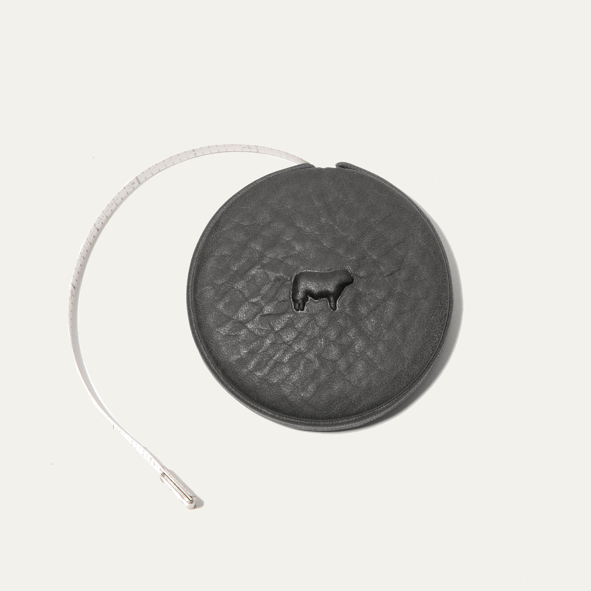Charcoal gray round leather tape measure case with an embossed cow logo, white cloth measuring tape partially extended to the left showing ruler markings.