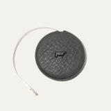 Charcoal gray round leather tape measure case with an embossed cow logo, white cloth measuring tape partially extended to the left showing ruler markings.