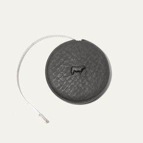 Charcoal gray round leather tape measure case with an embossed cow logo, white cloth measuring tape partially extended to the left showing ruler markings.