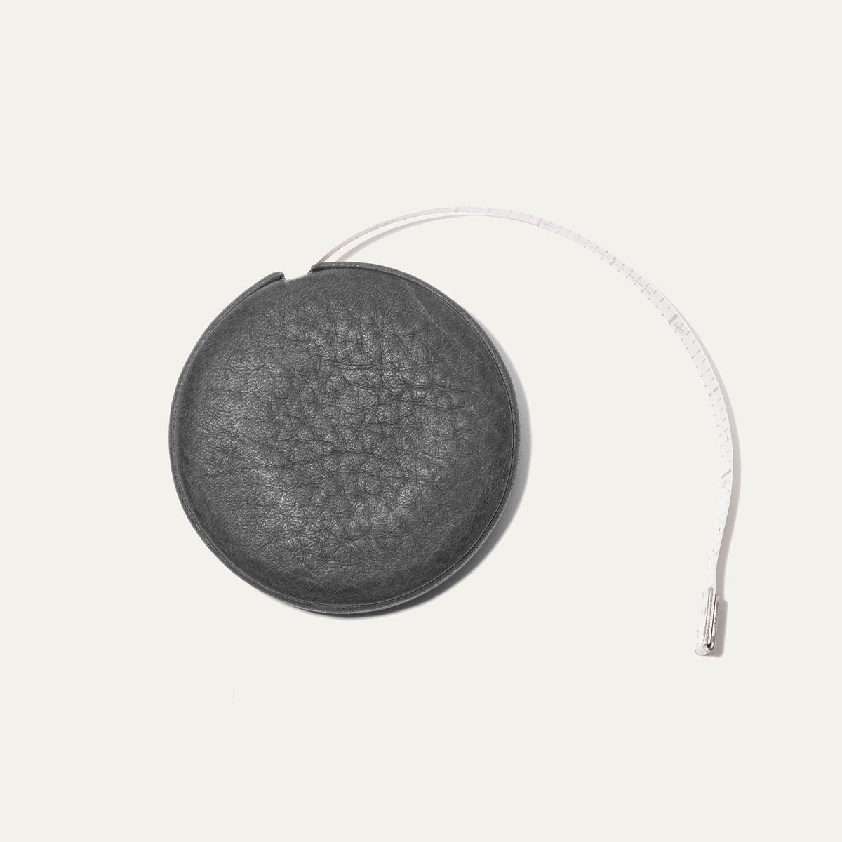 Charcoal gray round leather tape measure case with an embossed cow logo, white cloth measuring tape extended further in an arc showing ruler markings.