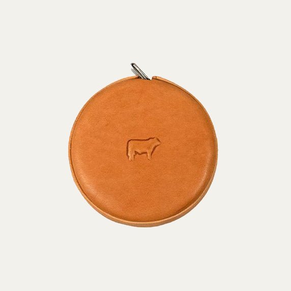 Cognac tan round leather tape measure case with embossed cow logo, front view showing a metal loop attachment at the top.