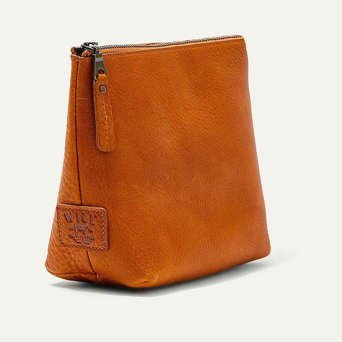 Tan leather zip pouch shown from a three-quarter angle, highlighting the trapezoid shape, zipper pull, and WILL leather patch