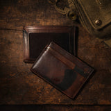 Lifestyle photo of blue and brown camo leather card cases resting on a wood table beside a canvas military jacket