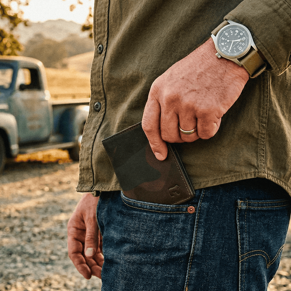 Lifestyle photo of a man's hand pulling the green camo Military Front Pocket wallet from his jeans back pocket, vintage pickup truck in the background.