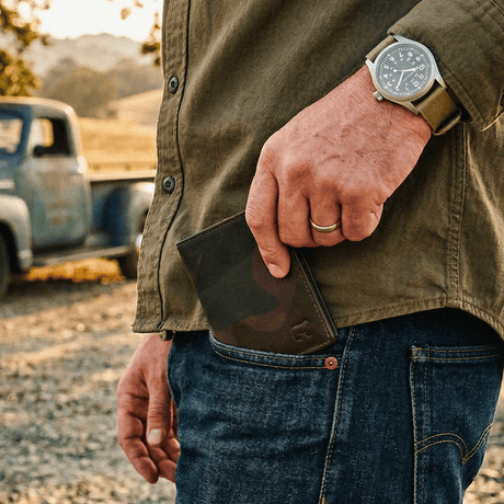 Lifestyle photo of a man's hand pulling the green camo Military Front Pocket wallet from his jeans back pocket, vintage pickup truck in the background.