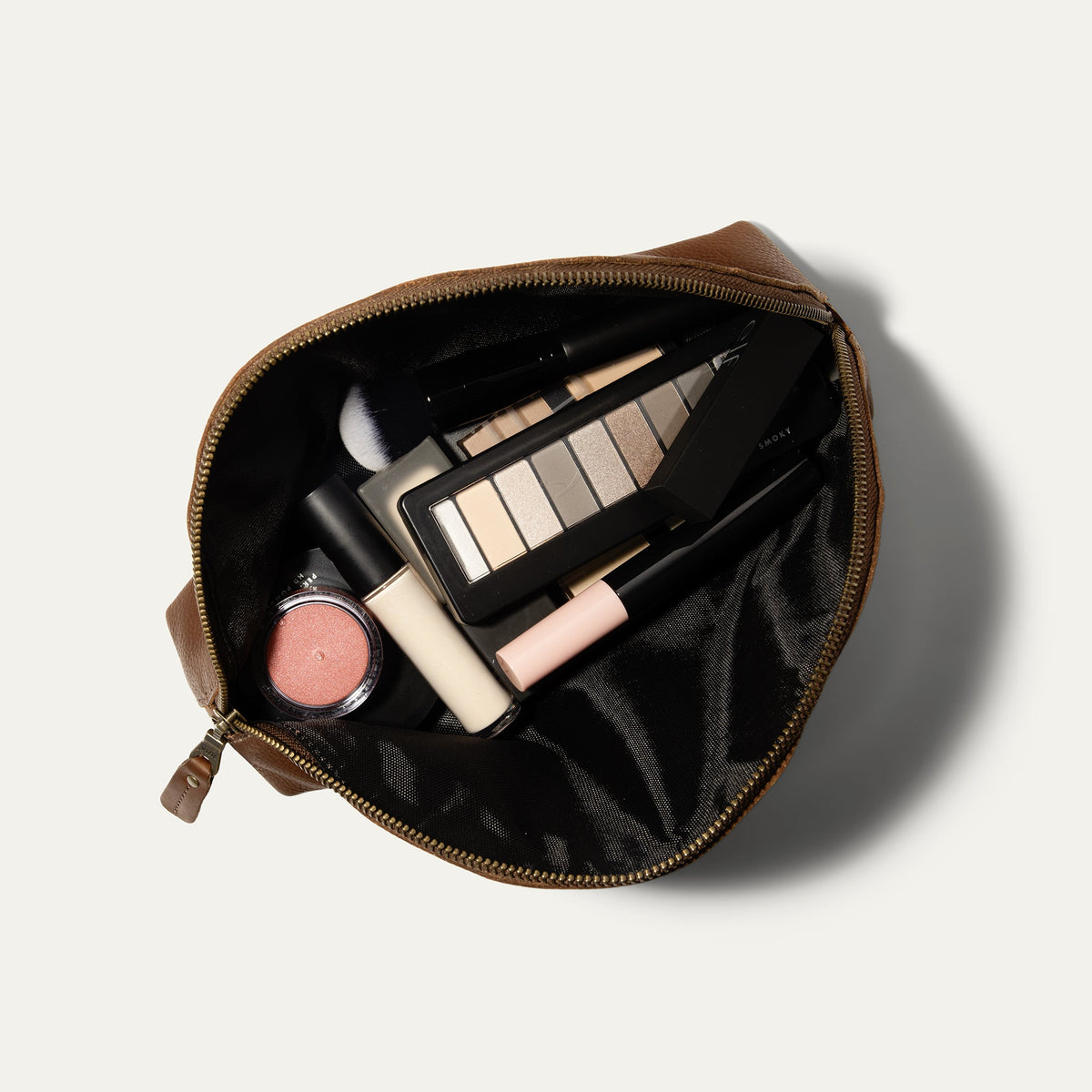 Overhead view of the cognac Zip Cosmetic bag open, unzipped to show the black mesh interior filled with eyeshadow palette, blush, concealer, lipstick, and a makeup brush.