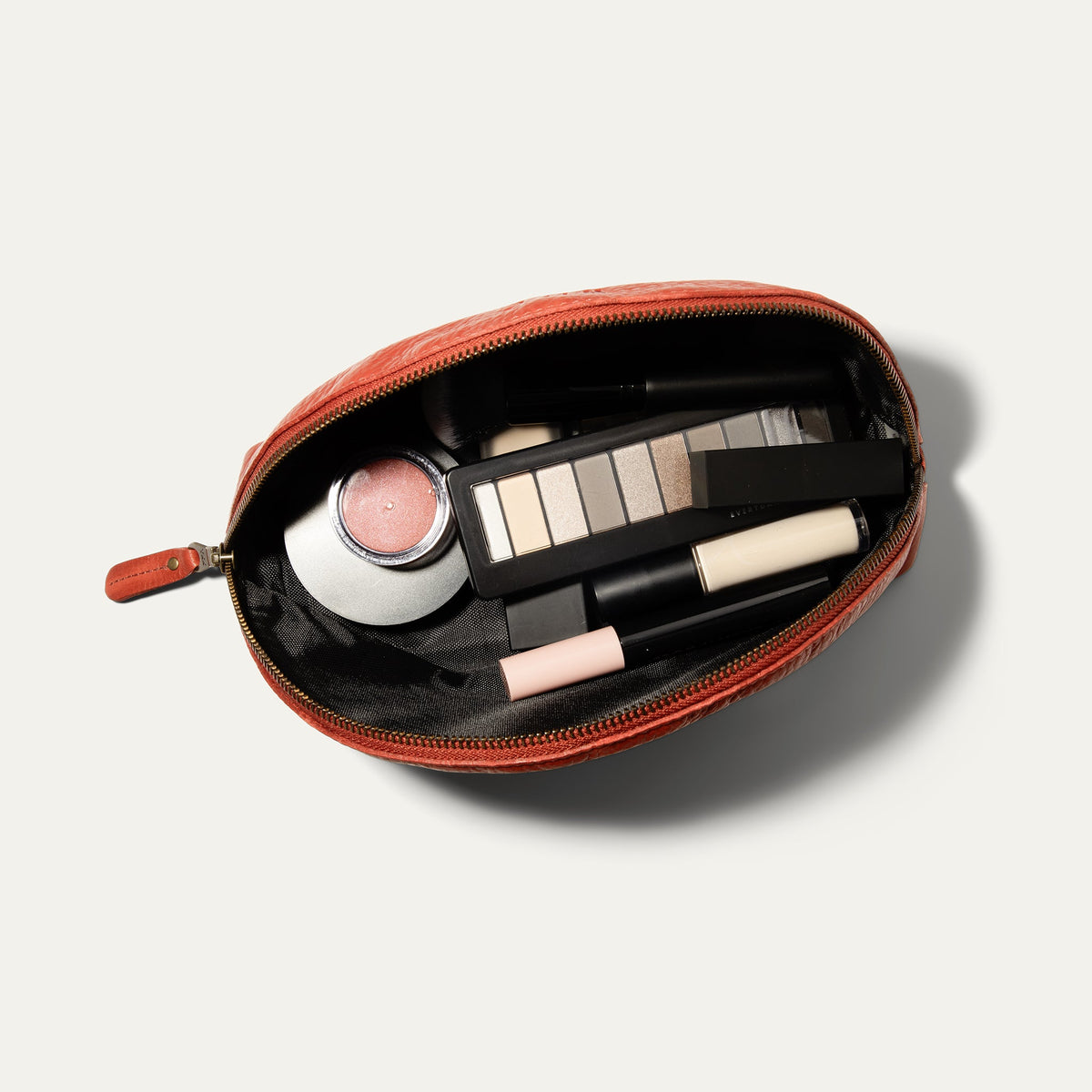 Overhead view of the red Zip Cosmetic bag open, unzipped to show the black mesh interior filled with a makeup brush, blush, concealer, eyeshadow palette, and lip products.