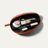Overhead view of the red Zip Cosmetic bag open, unzipped to show the black mesh interior filled with a makeup brush, blush, concealer, eyeshadow palette, and lip products.