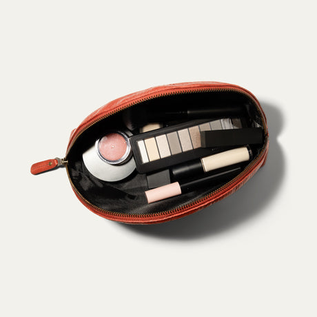 Overhead view of the red Zip Cosmetic bag open, unzipped to show the black mesh interior filled with a makeup brush, blush, concealer, eyeshadow palette, and lip products.