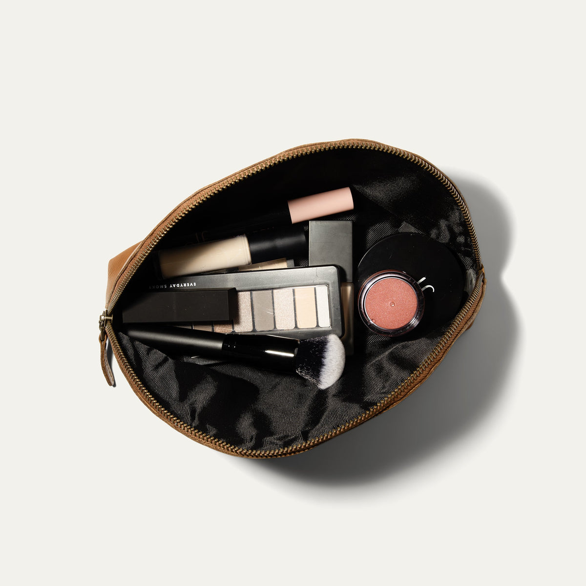 Overhead view of the tan Zip Cosmetic bag open, unzipped to show the black mesh interior filled with concealer, eyeshadow palette, lip balm, and a makeup brush.